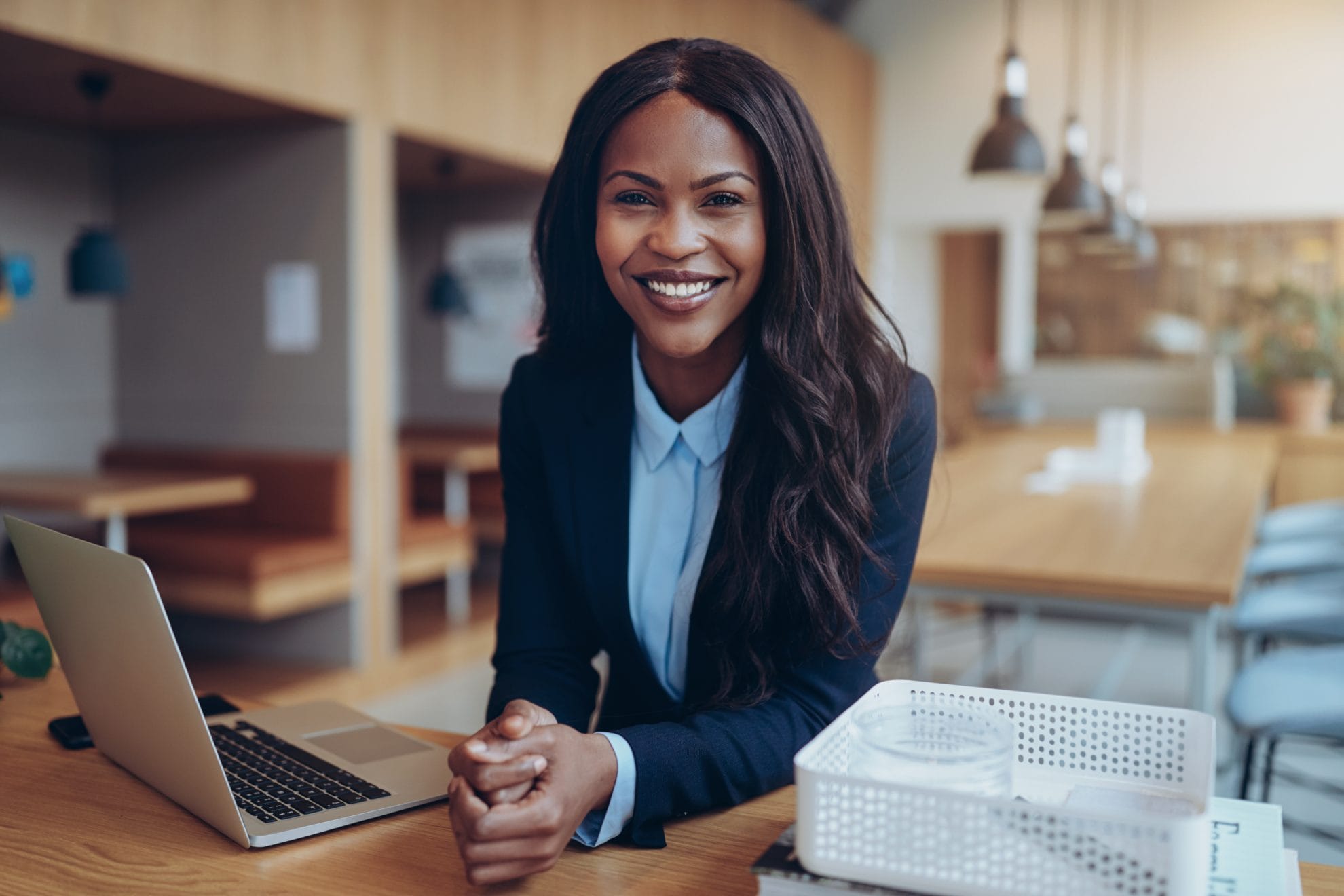  A smiling black businesswoman leaning on a desk and looking at the camera while working on a laptop.